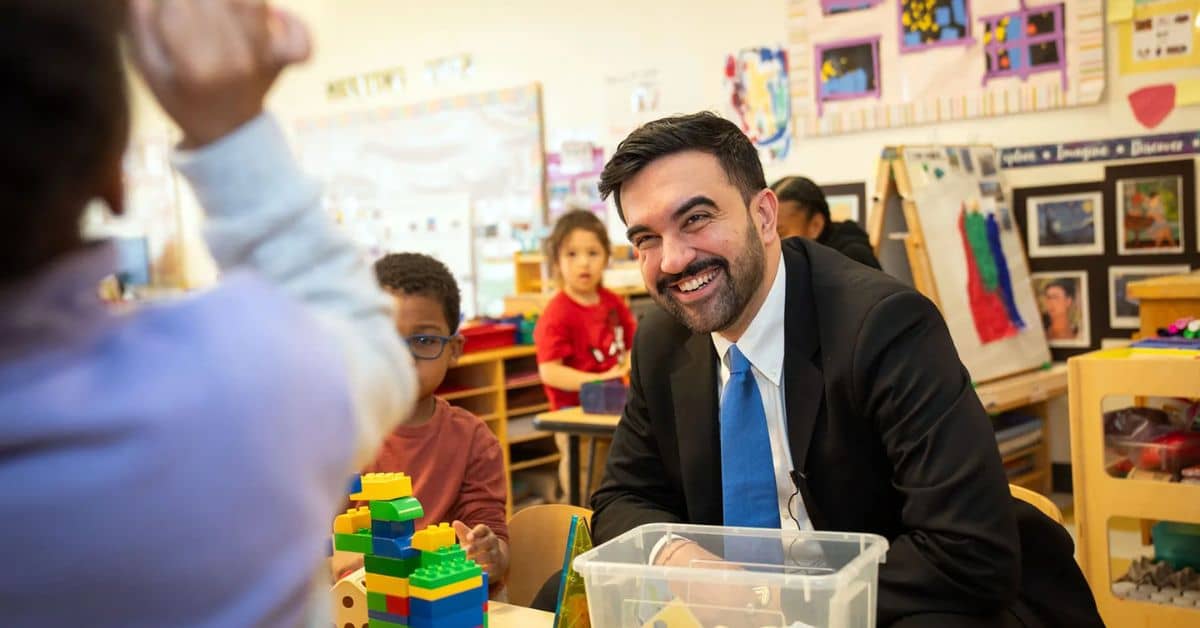 Man in black suit with a blue tie sitting at a low children's table with blocks on the table, he's smiling at a child across the table