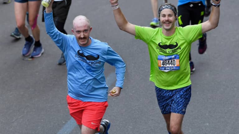 Two people running, smiling with their arms overhead, wearing bright shirts with mustaches on them