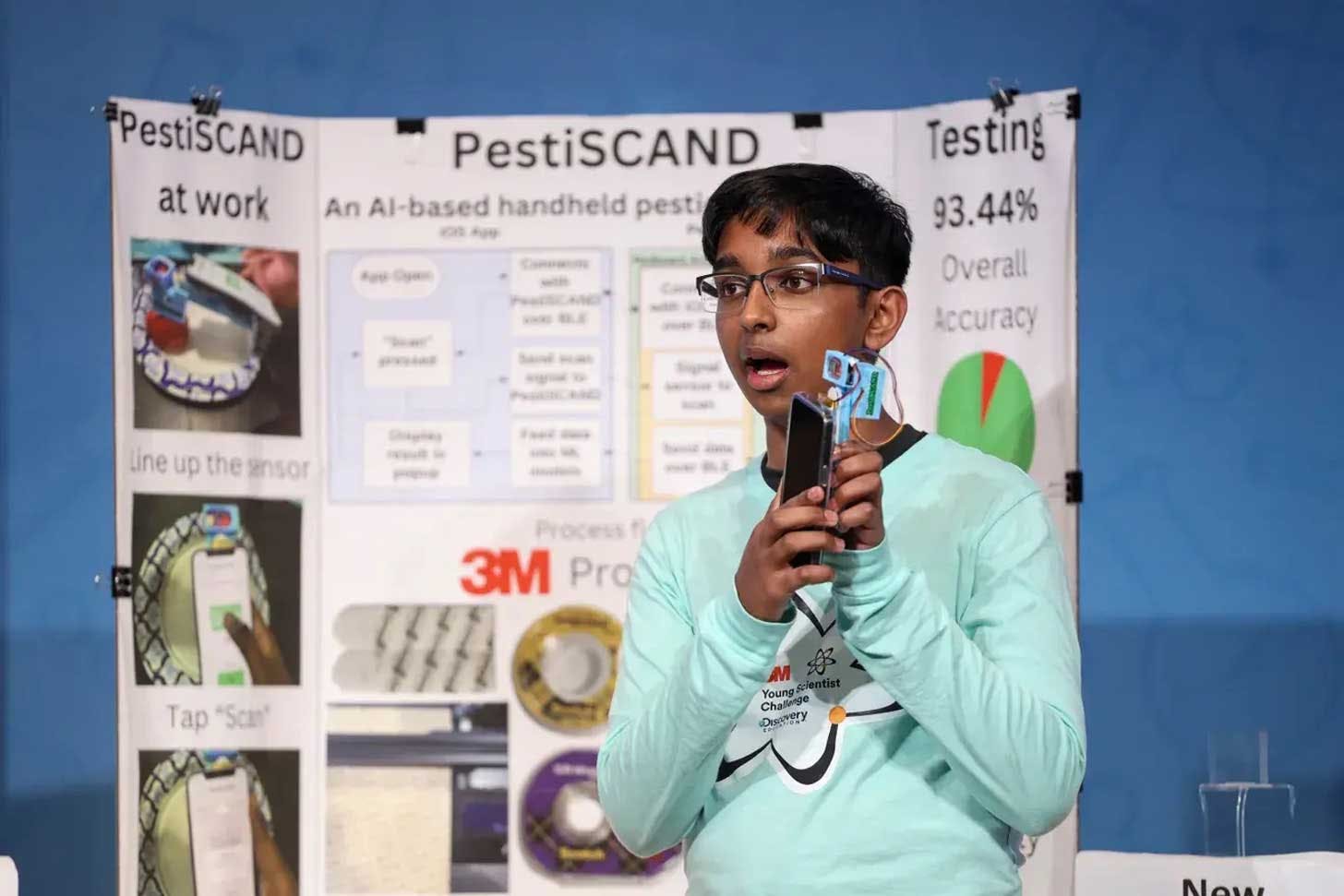 A teen in a light teal long sleeve shirt holding a hand-held scanner device in front of a tri-fold presentation board