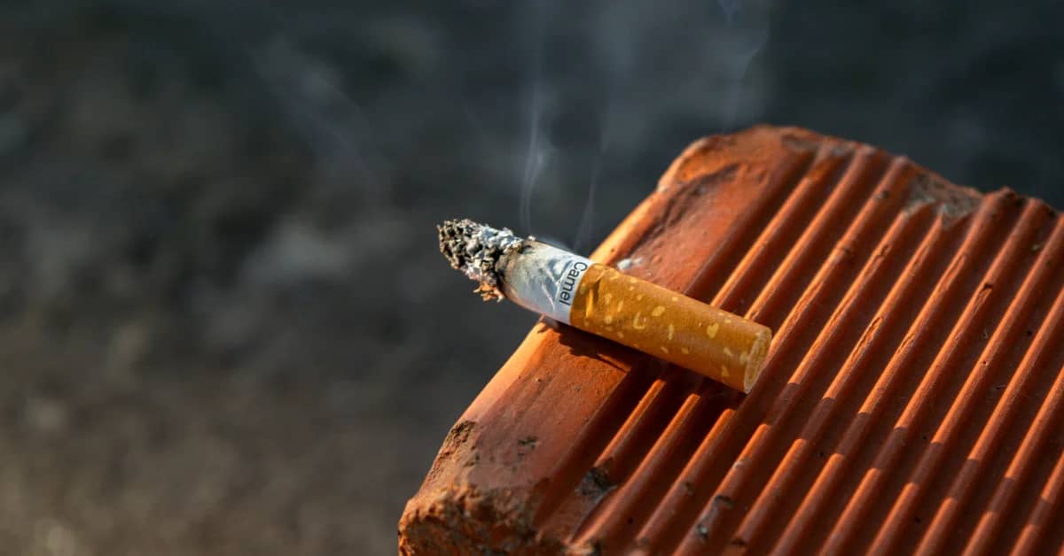 A lit cigarette rests on stacked terracotta roof tiles against a dark, blurred background. Smoke rises from the cigarette's tip.