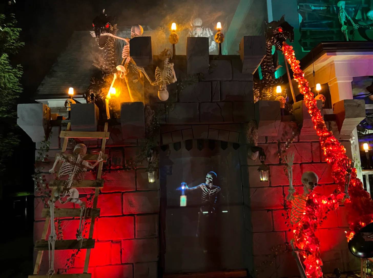 A castle facade decorated for Halloween displays skeleton figures holding torches and lanterns, red LED garland vines, and a glowing skeleton figure in the center gateway. Black castle walls are illuminated with red and amber lighting.