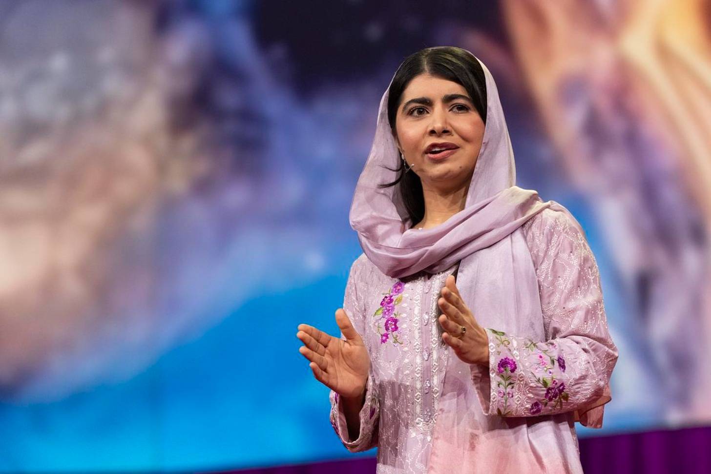 Malala wears a light pink, floral embroidered dress, matching head scarf, stands on stage wearing a small microphone in front of a colorful background