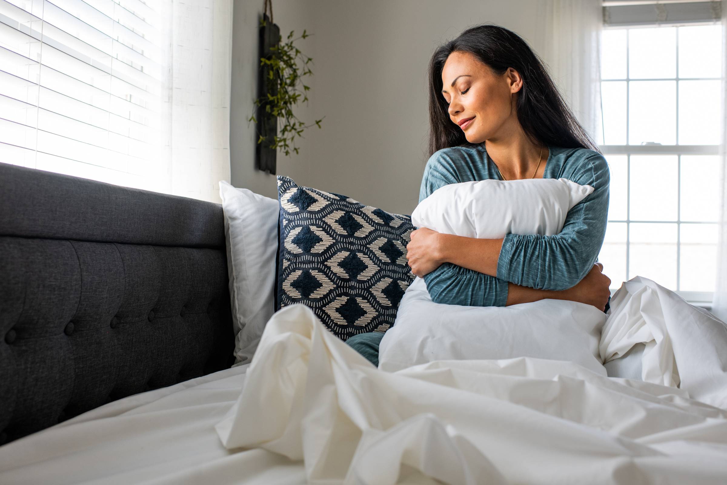 A person wearing a teal long-sleeved shirt sitting upright in bed hugging a pillow with white sheets on the bed
