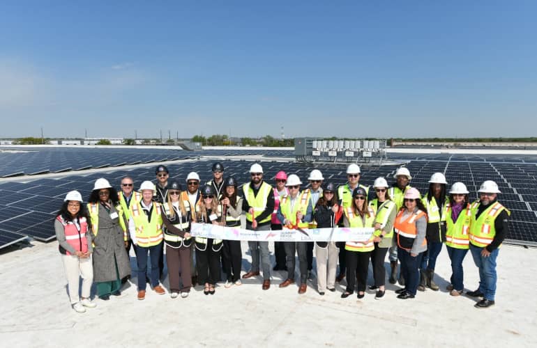 A group of about 25 people in yellow vests and hard hats standing on top of a building covered in solar panels