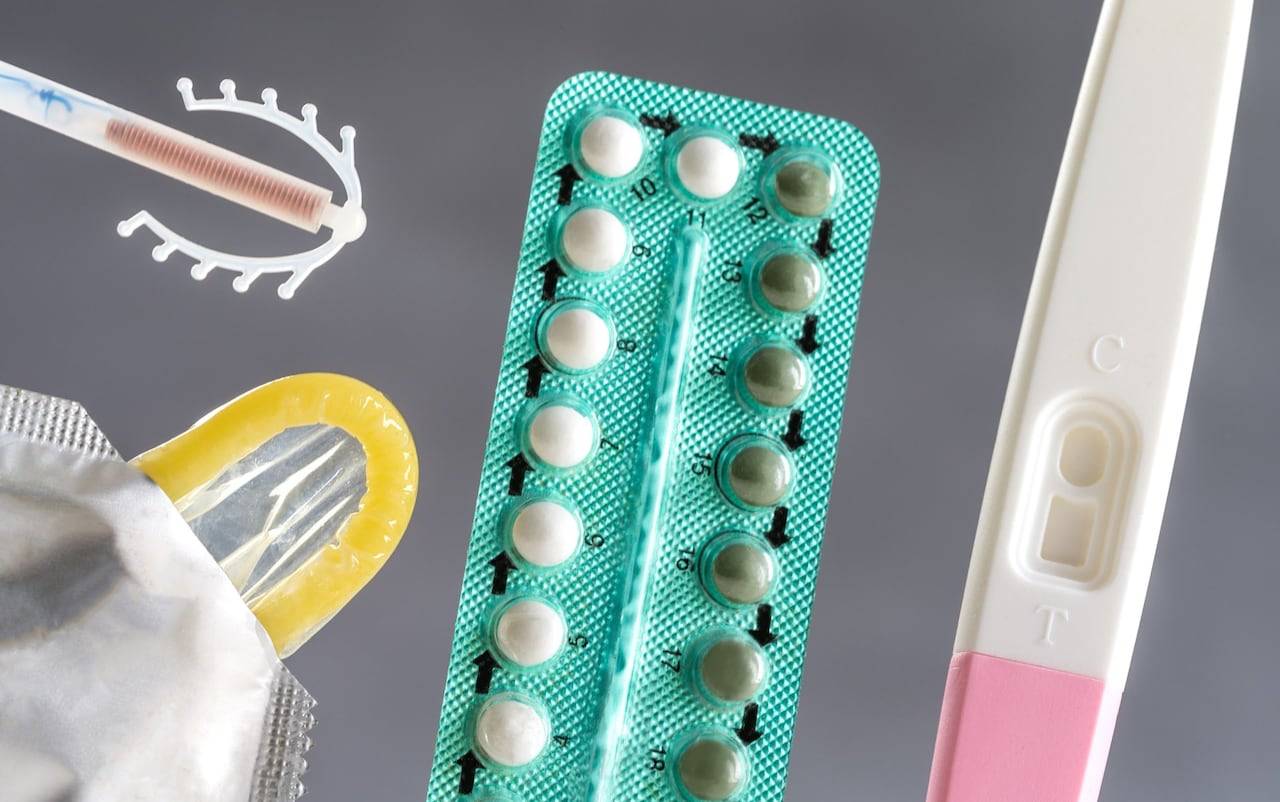 A turquoise blister pack of oral contraceptive pills, a yellow condom wrapper, a pregnancy test showing a pink result line, and a toothbrush arranged on a gray background.