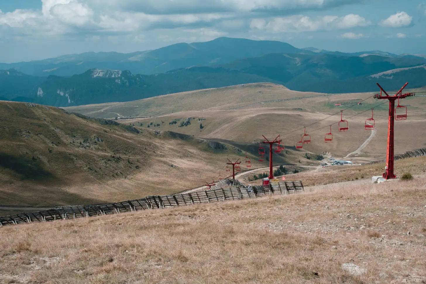 Red ski lift towers with empty chairs extend across a dry, rolling hillside, with forested mountains visible in the background under a partly cloudy sky.