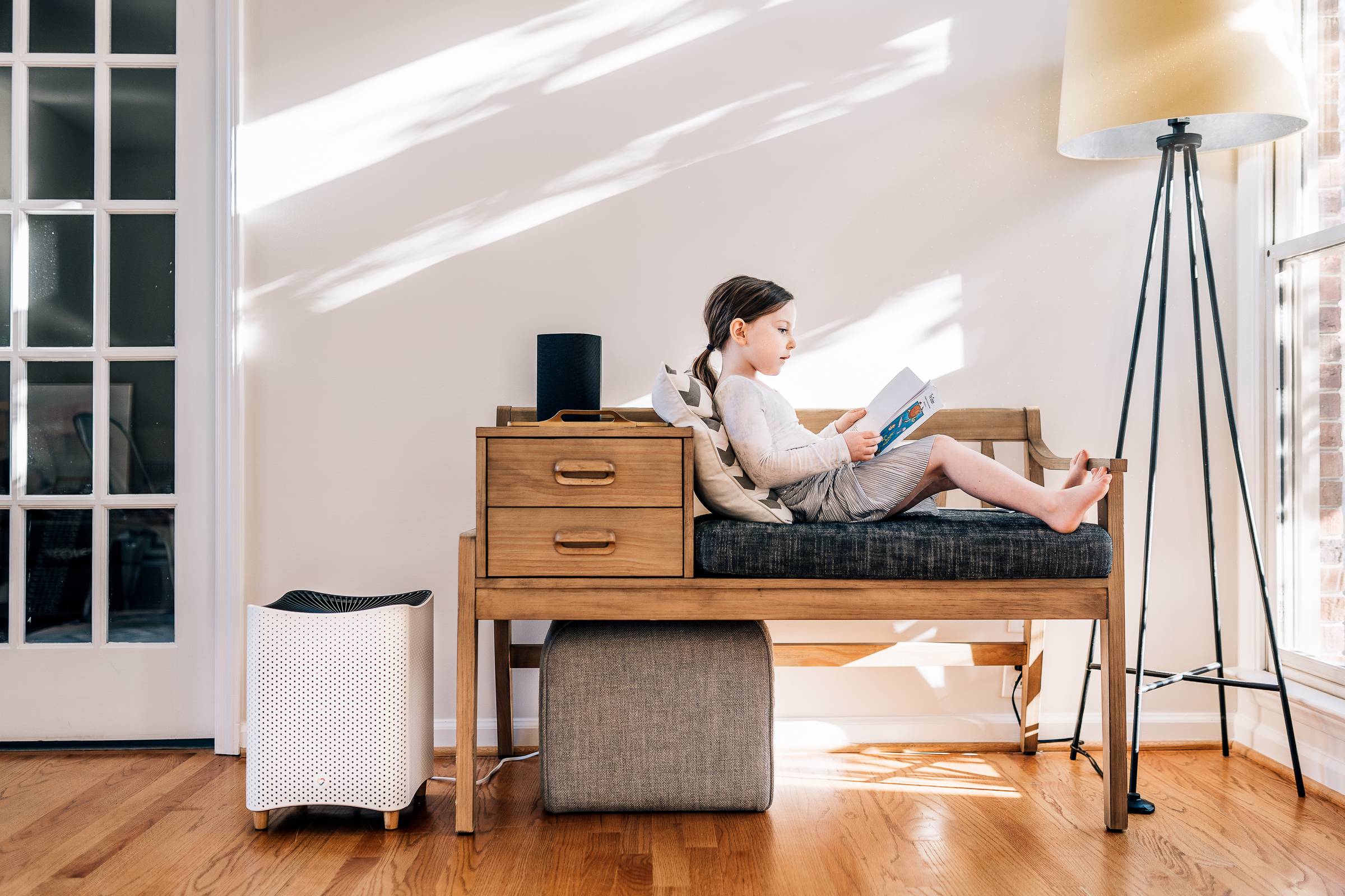 A young girl sitting on a padded bench reading a book next to a Mila Cares air purifier