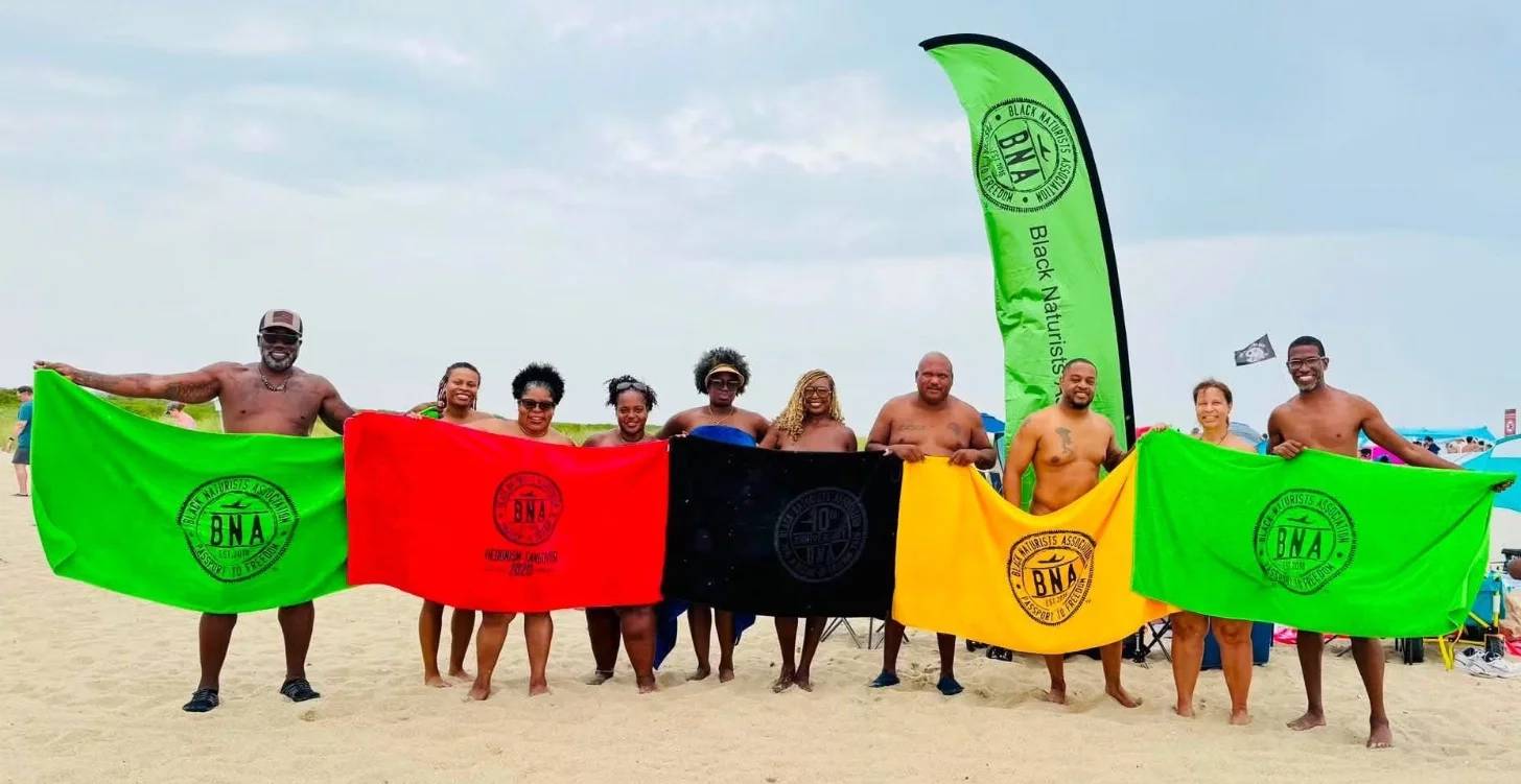 Around 10 people on a beach holding up green, red, black, yellow towels with a black symbol on them.