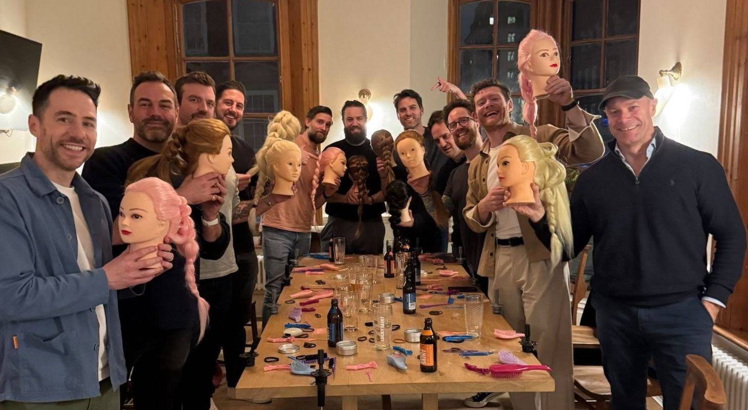 A group of more than a dozen dads of varying ages smile as they gather around a long table in a pub and hold up mannequin heads with different braids and styles