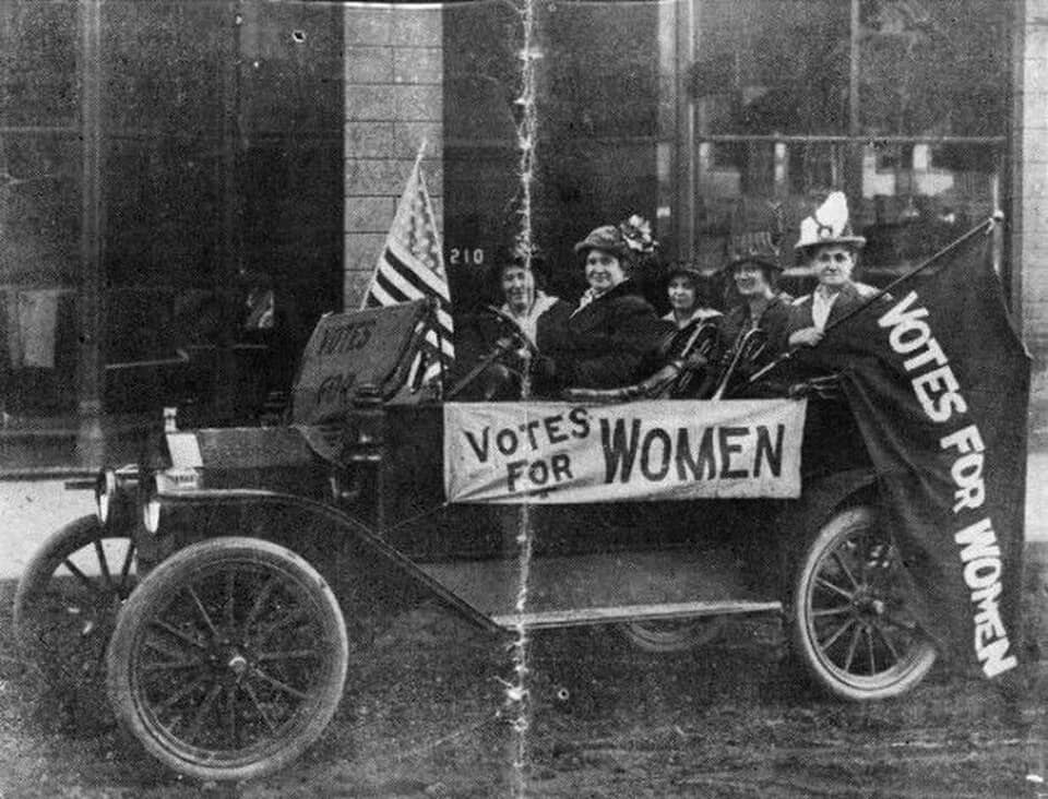 A black and white photo of five women in an old car, a banner that says "Votes for Women" is hung from the side of the car