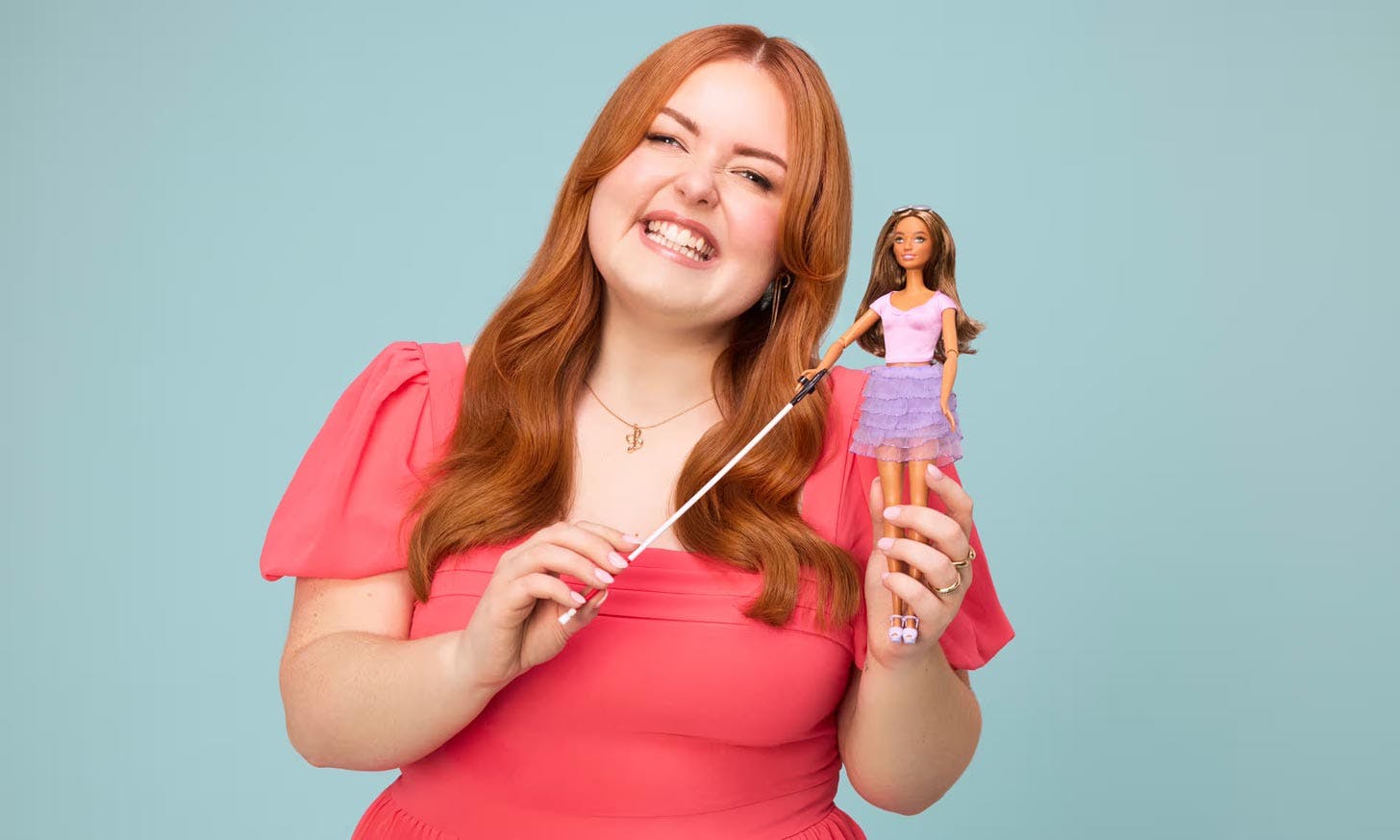 A person in a pink dress smiles holding Blind Barbie on a light blue backdrop