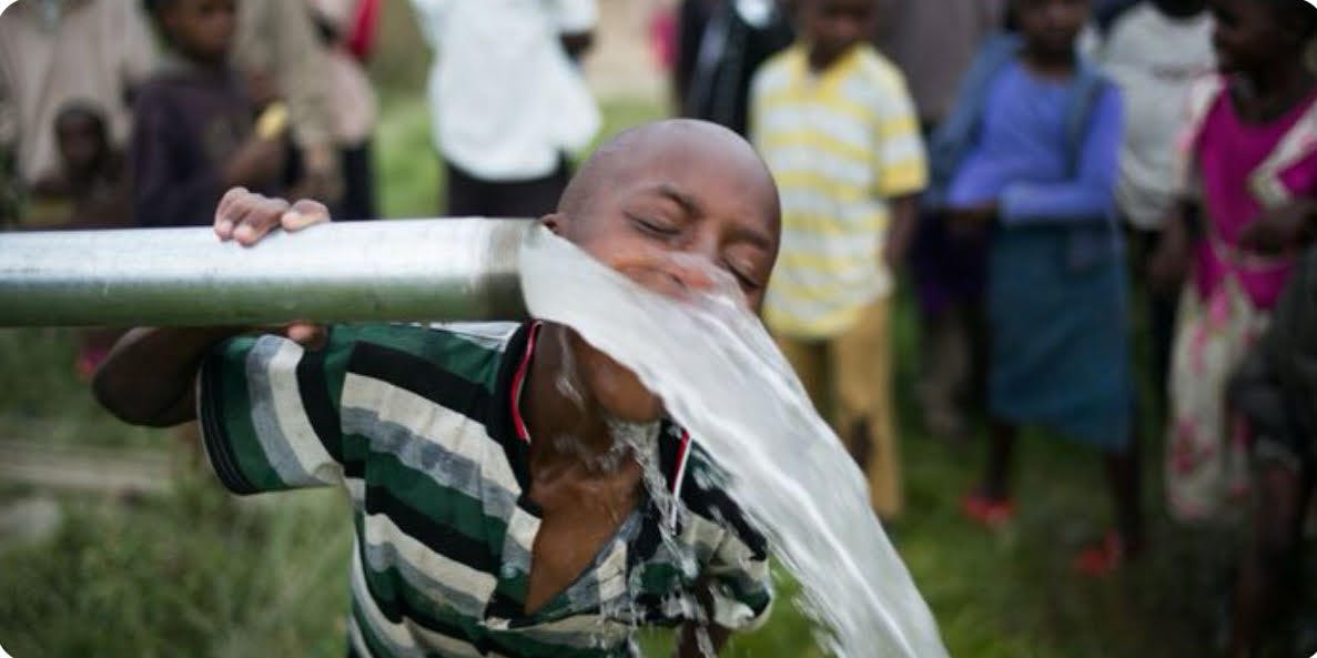 An older child wearing a striped shirt, they're putting their face in a stream of water coming out of a metal pipe