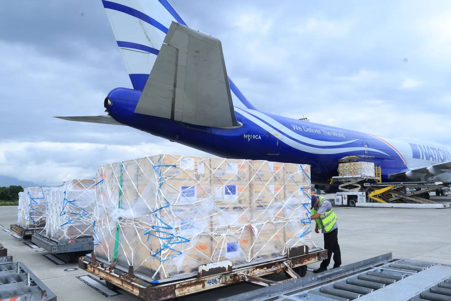 A cargo airplane with blue and white livery is loaded with wrapped pallets of freight. A worker in a yellow safety vest stands near the cargo on the tarmac. The aircraft displays "We Deliver The World" text on its fuselage and registration number N919CA.