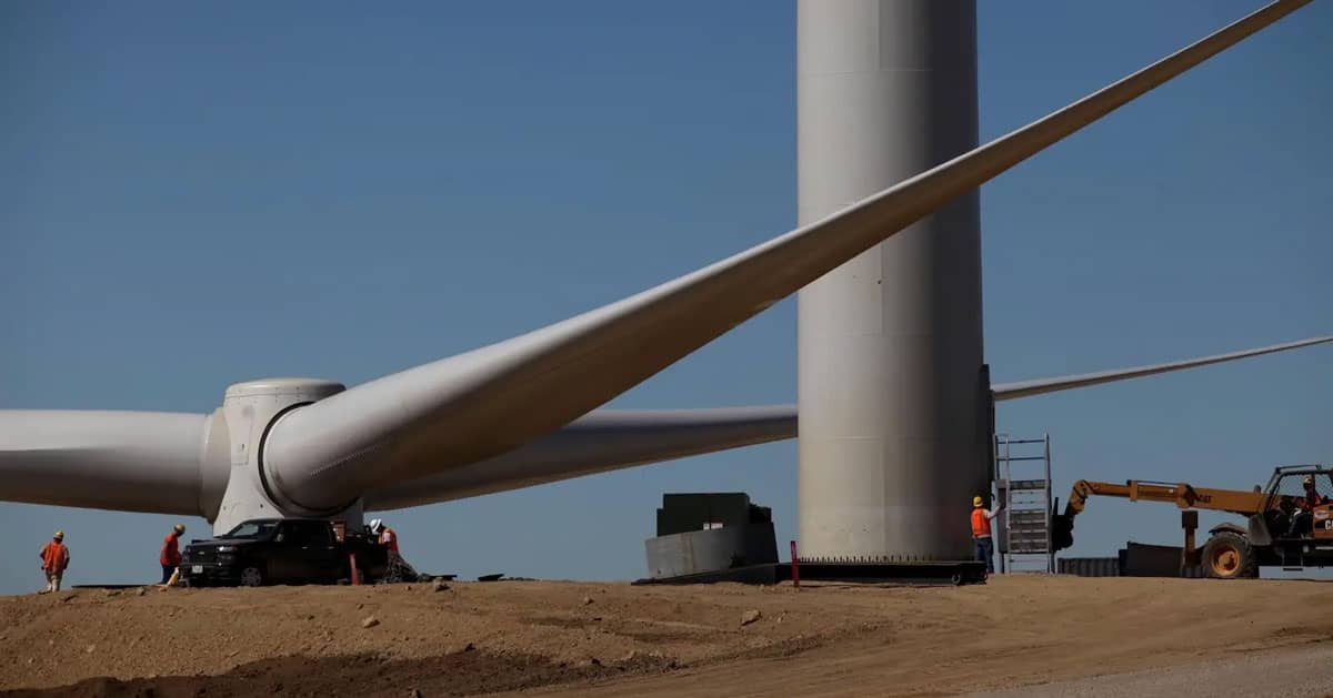 Cars and workers at the base of a wind turbine with the turbine blades sitting on the ground waiting for installation