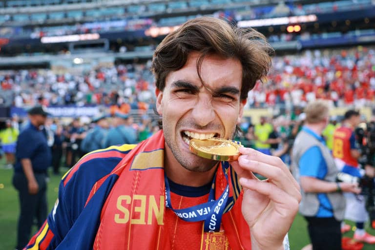 A person looking at the camera, biting into a round gold medal on a blue ribbon