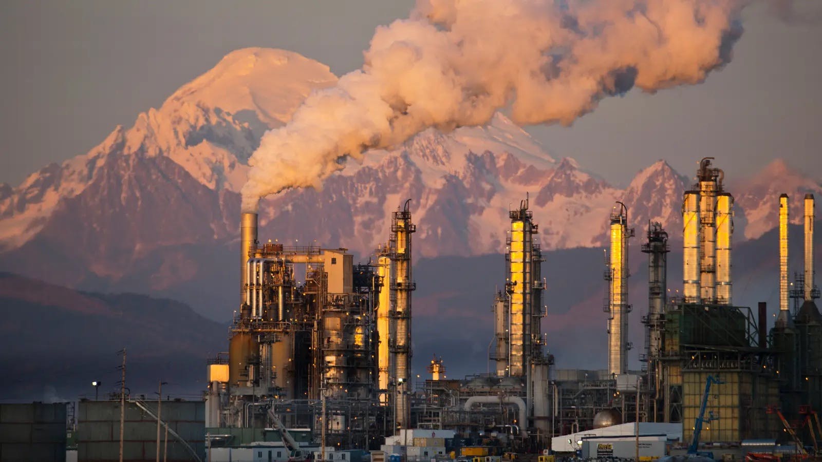 An oil refinery with smoke coming out of one chimney, a snow-capped mountain is visible in the background