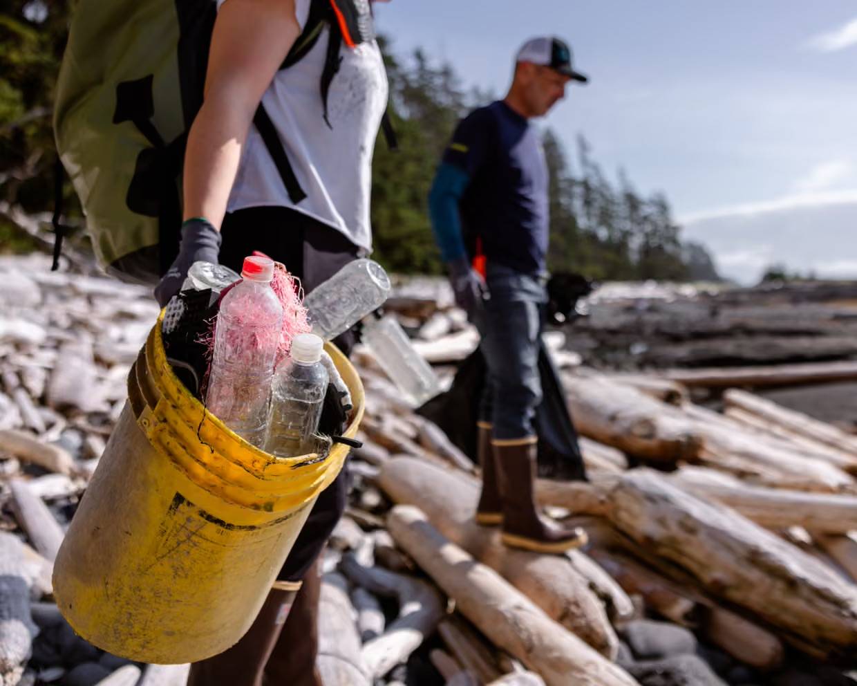 A volunteer in a yellow bucket holds plastic water bottles wrapped in pink netting during a beach cleanup, with other volunteers and driftwood visible in the background.