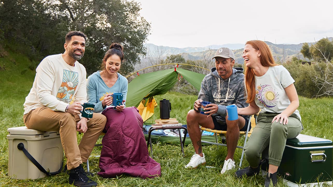 Four people wearing Life is Good shirts holding mugs and sitting on camping chairs and coolers with a tent and mountains in the background