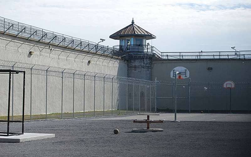 Prison recreation yard with basketball hoop, high concrete walls topped with razor wire fencing, and a guard tower with conical roof.