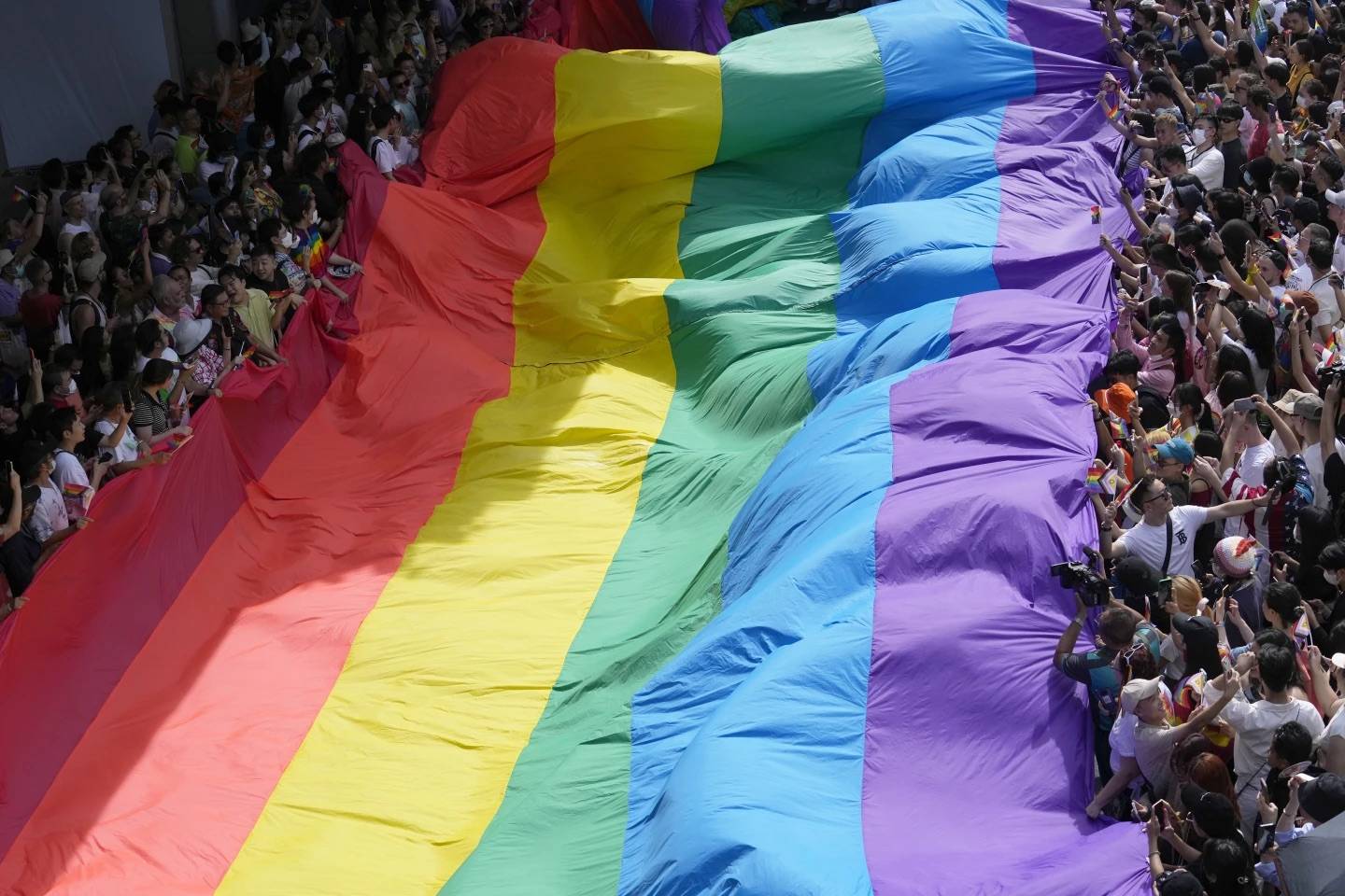 A massive rainbow pride flag held on either side by a crowd of people