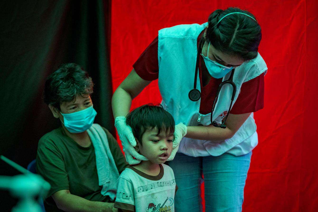 A smaller child gets their jawline examined by a medical provider while their caregiver sits behind them