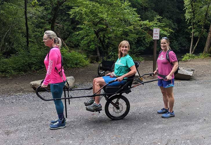 Two people holding the front and back end of an accessible hiking bike with a person sitting in the middle in a chair