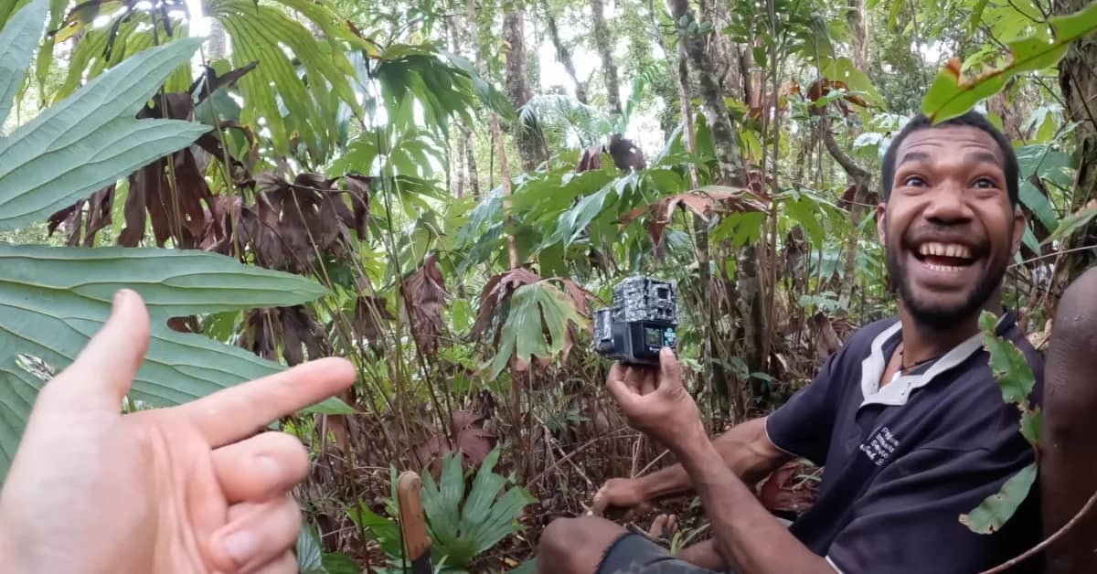 A person holding a go-pro-style camera smiling at someone standing behind him, they are in a dense wooded area