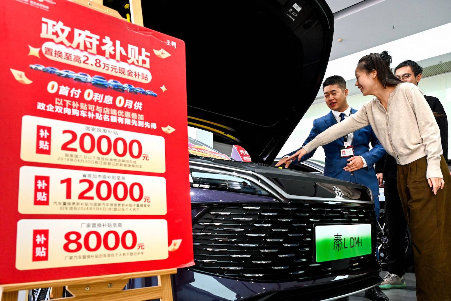 A salesman in a blue jacket shows a black car with green LED display to a woman in a beige sweater at an auto showroom. A red promotional sign displaying government subsidies of 20000元, 12000元, and 8000元 stands beside them.