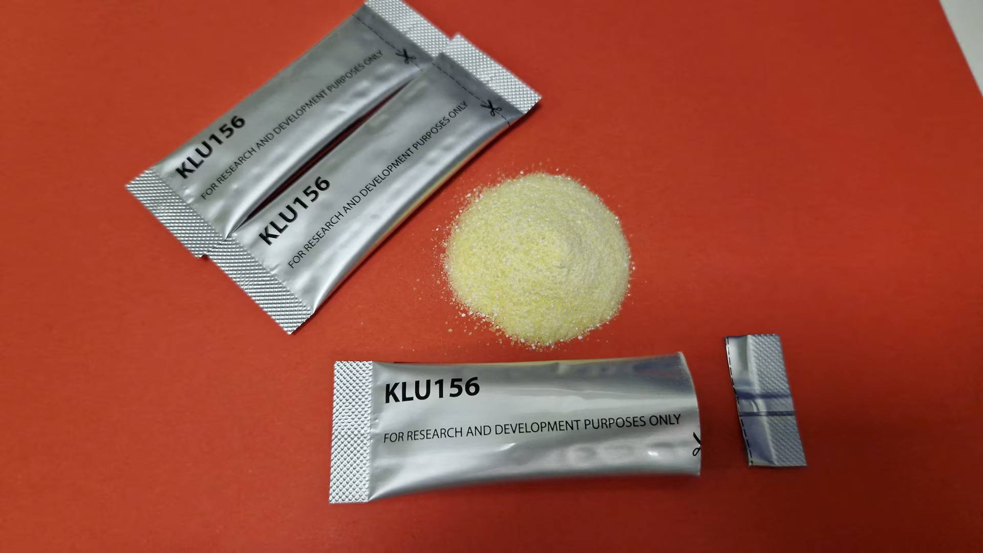 Three silver packets labeled "KLU156", one is open with a yellowy powder on a red background