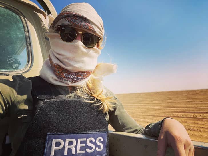 Jane in the back of a truck wearing a scarf over her head and face, sunglasses, and a press vest