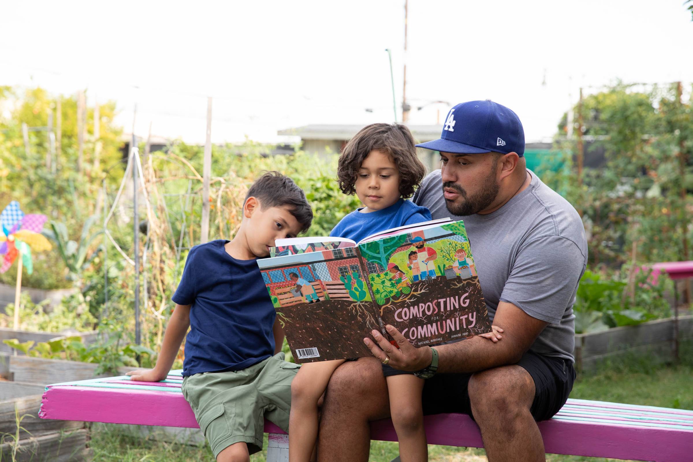 A person in a blue baseball cap and grey tee with a child on his lap and another child sitting next to him reading a book titled "Composting for Community"