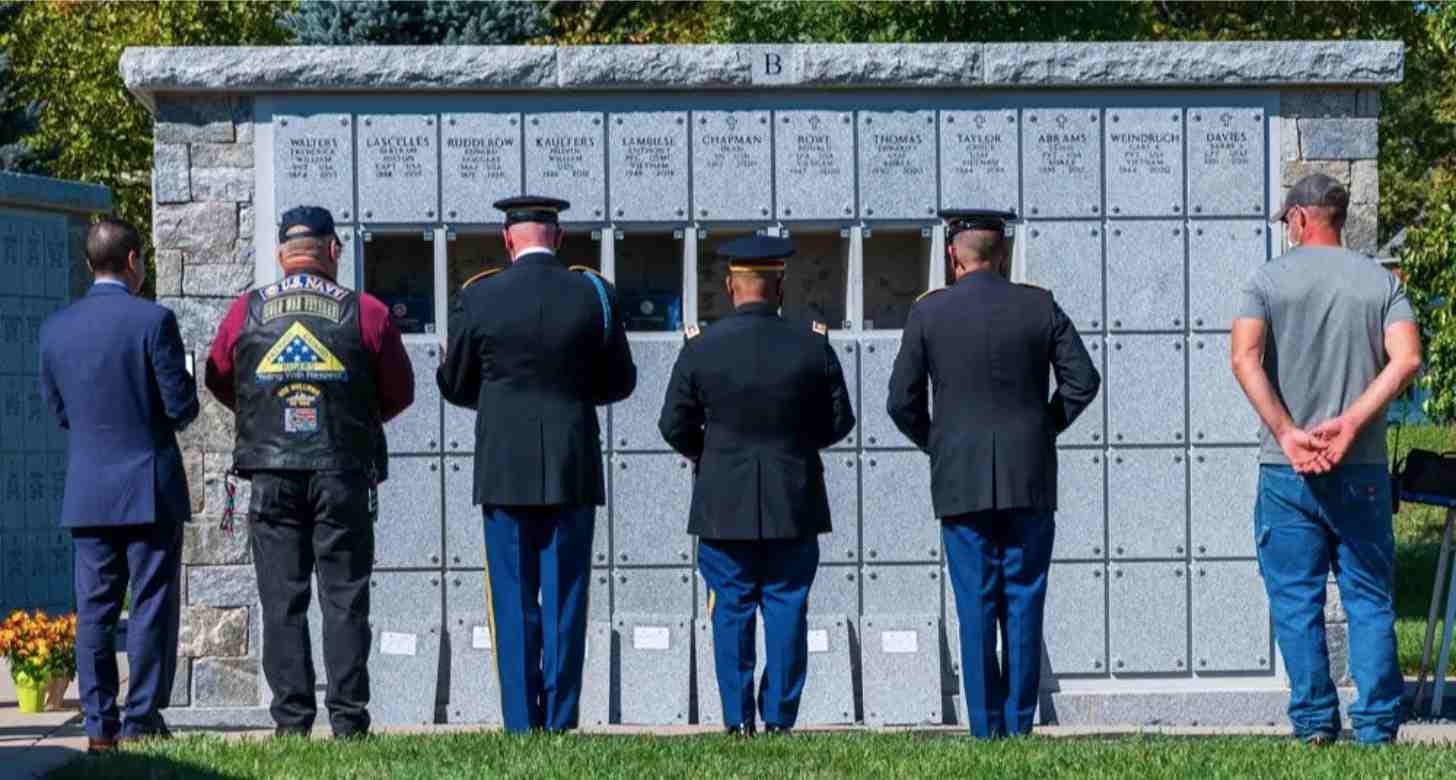 Six people standing in front of a stone wall in a cemetary, three people are in military uniforms, there are several compartments on the stone wall open while others are closed