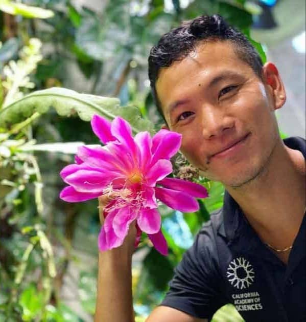 Tim Wong smiling at the camera next to a bright pink flower with greenery in the background