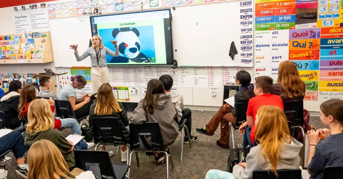 A teacher stands at the front of a French language classroom, gesturing toward a screen displaying a panda with the text "La musique." Students sit in chairs facing the teacher, with French grammar posters and vocabulary charts visible on the walls behind them.