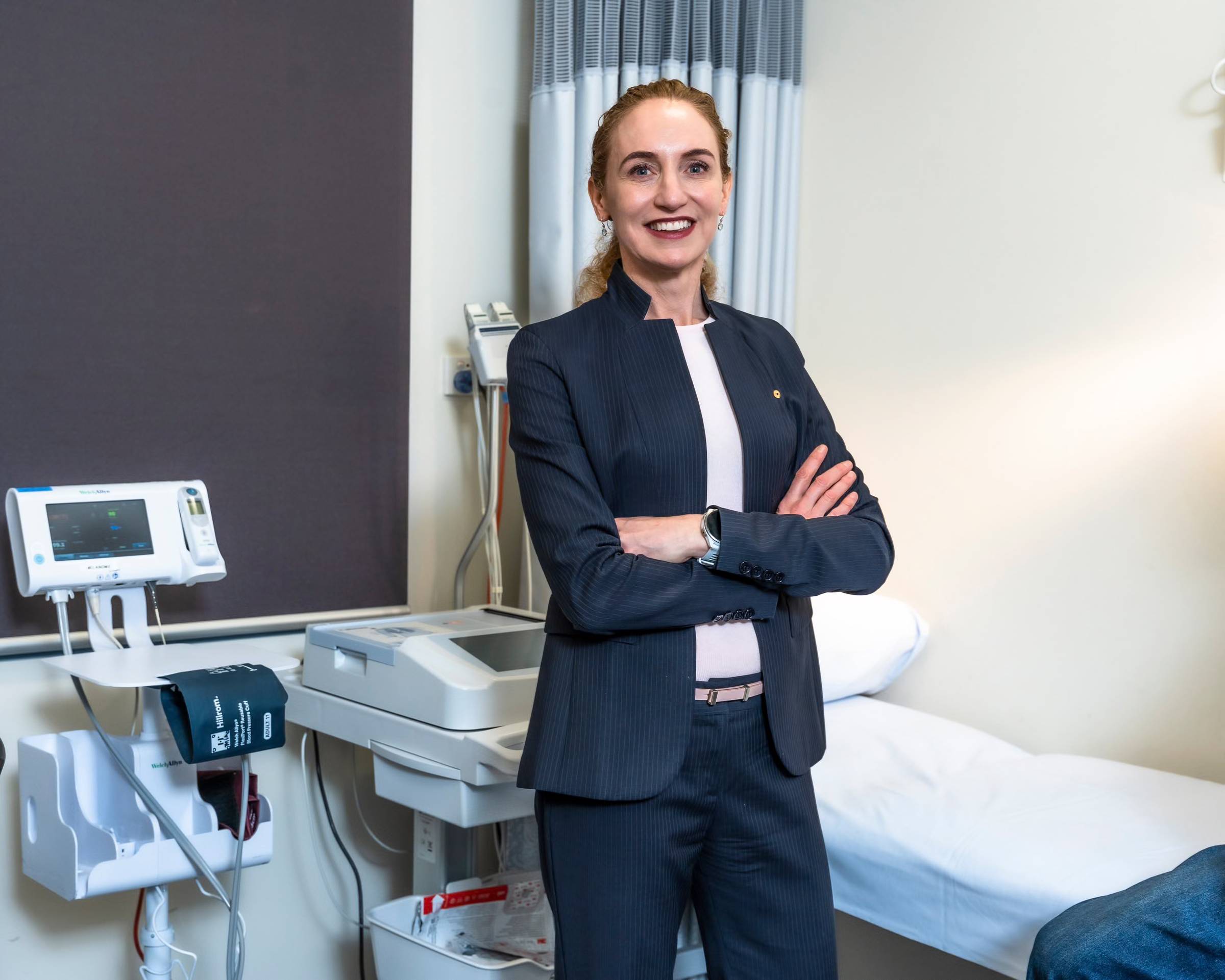 Woman in dark pinstriped business suit standing with arms crossed in a medical examination room, with diagnostic equipment visible on the counter and examination table behind her.