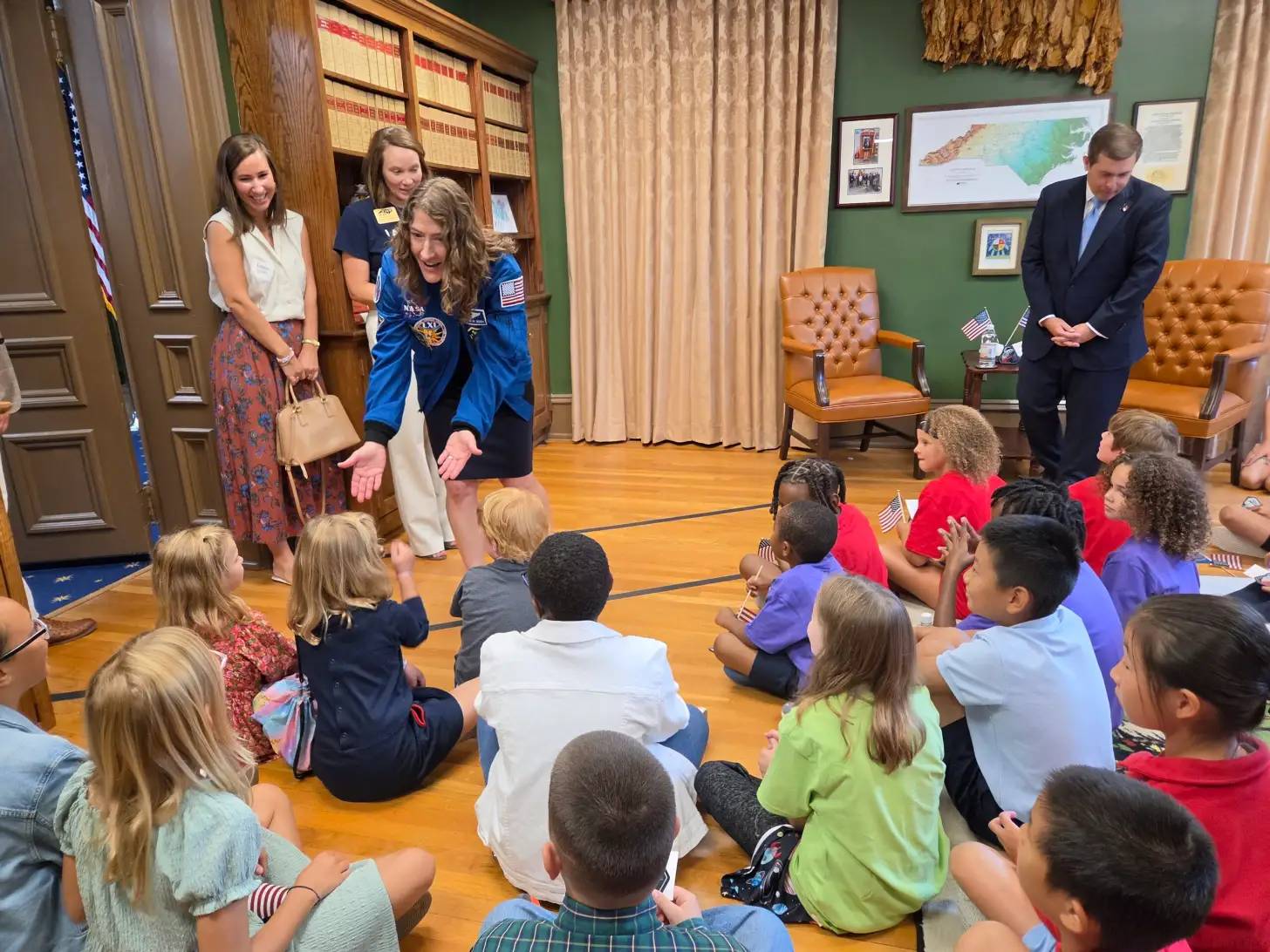 A NASA astronaut in a blue flight suit sits with a group of children in an official government office. Adults stand nearby, including a man in a dark suit and women in business attire.