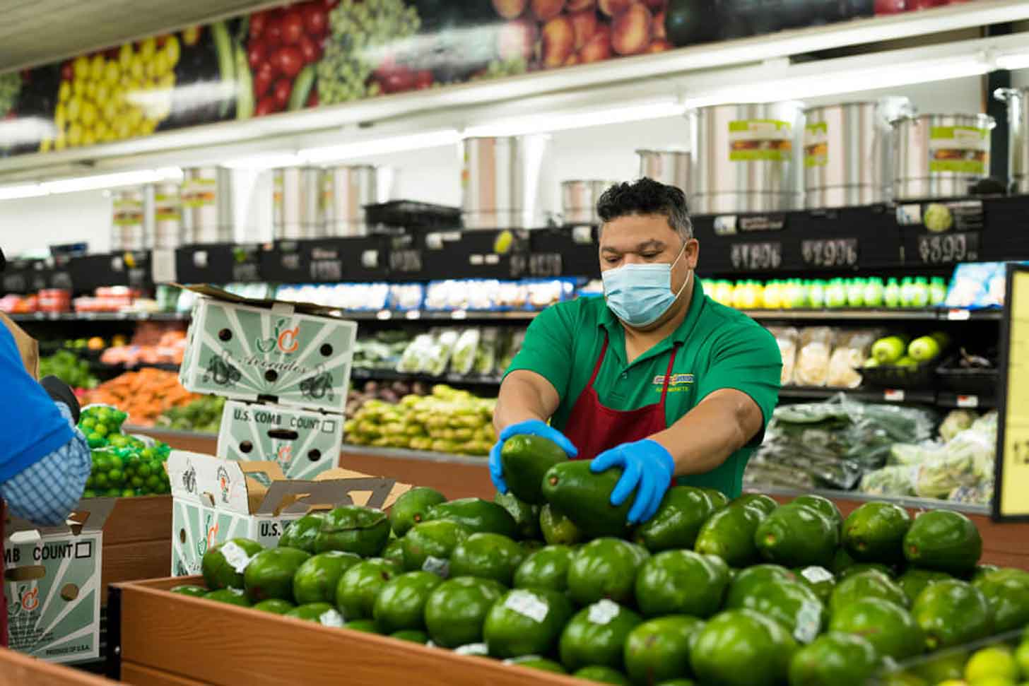 A worker in a blue mask, green shirt, and red apron putting produce on a display
