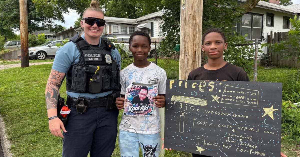 A police officer standing with two young boys holding signs for their lemonade stand