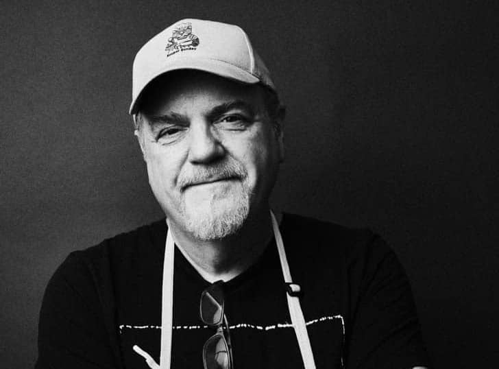 Black and white photo of a man in a hat, black shirt, and apron