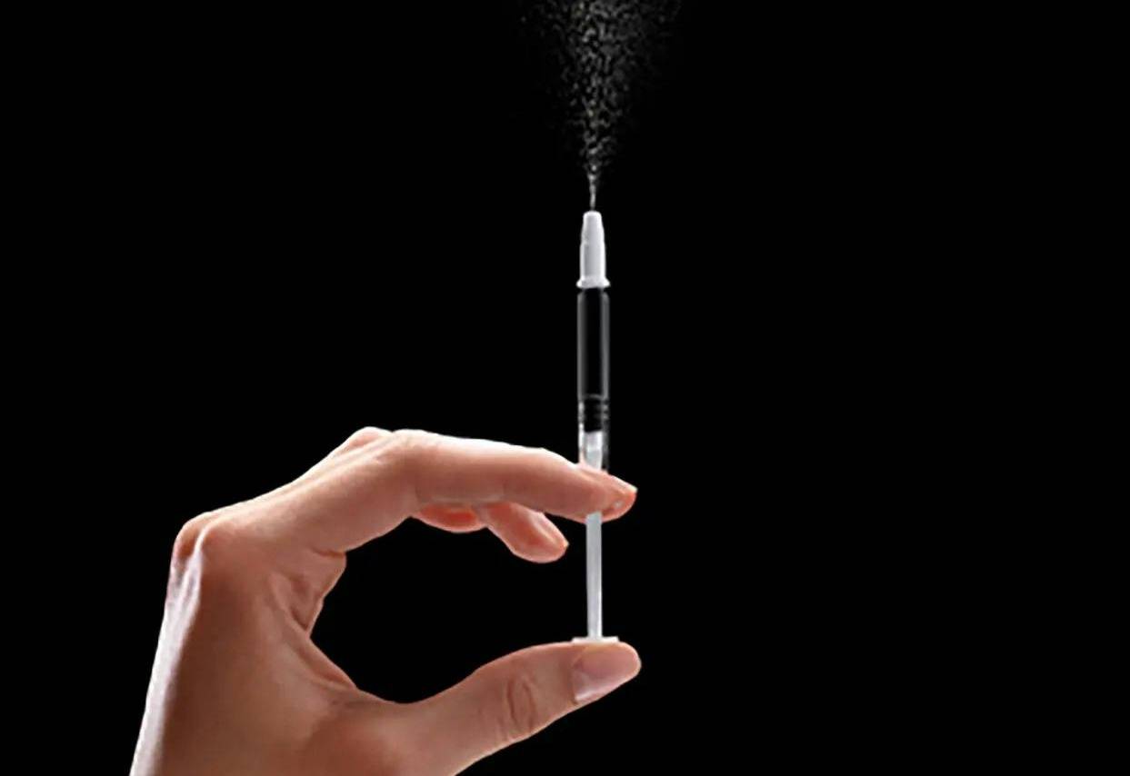 A hand holding a syringe-looking nasal sprayer on a completely black background, some liquid is misting out of the top of the syringe