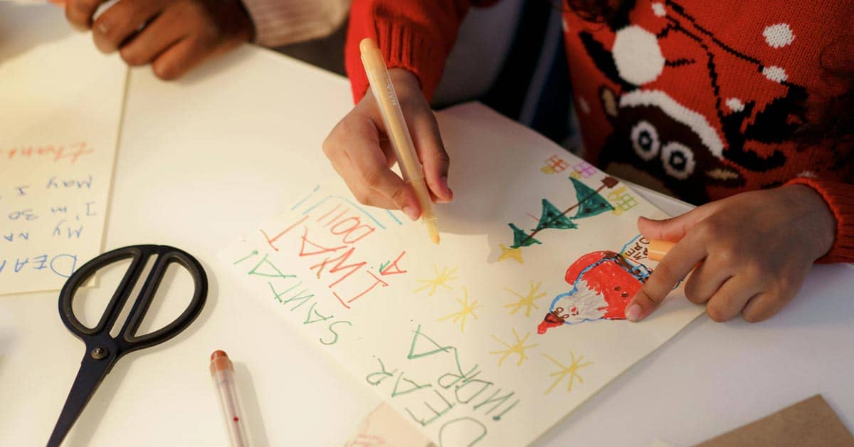 A child drawing a letter to santa on a white table, they're wearing a sweater with rudolph on it, the letter says "Dear santa, I want a doll!"