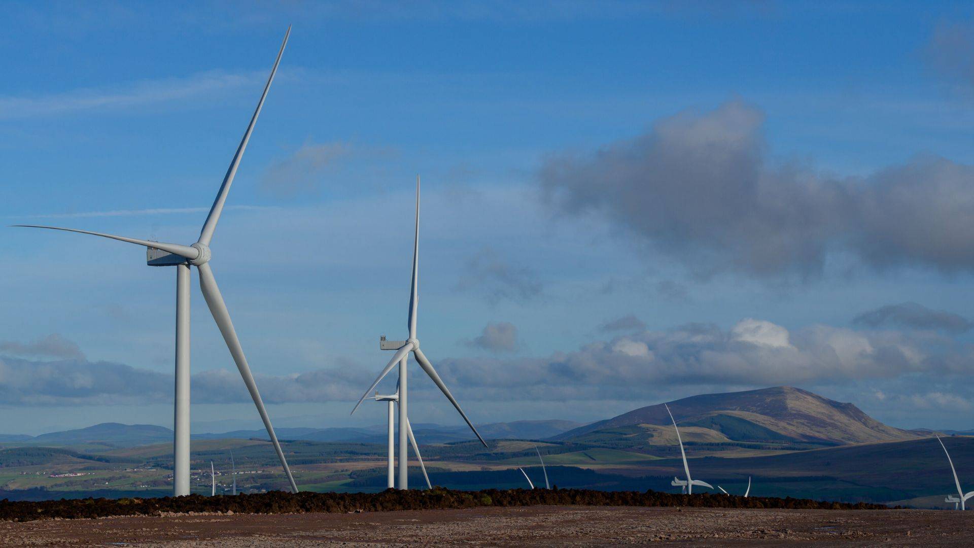 Several white wind turbines stand on hilly terrain with mountains visible in the distance under a partly cloudy sky.