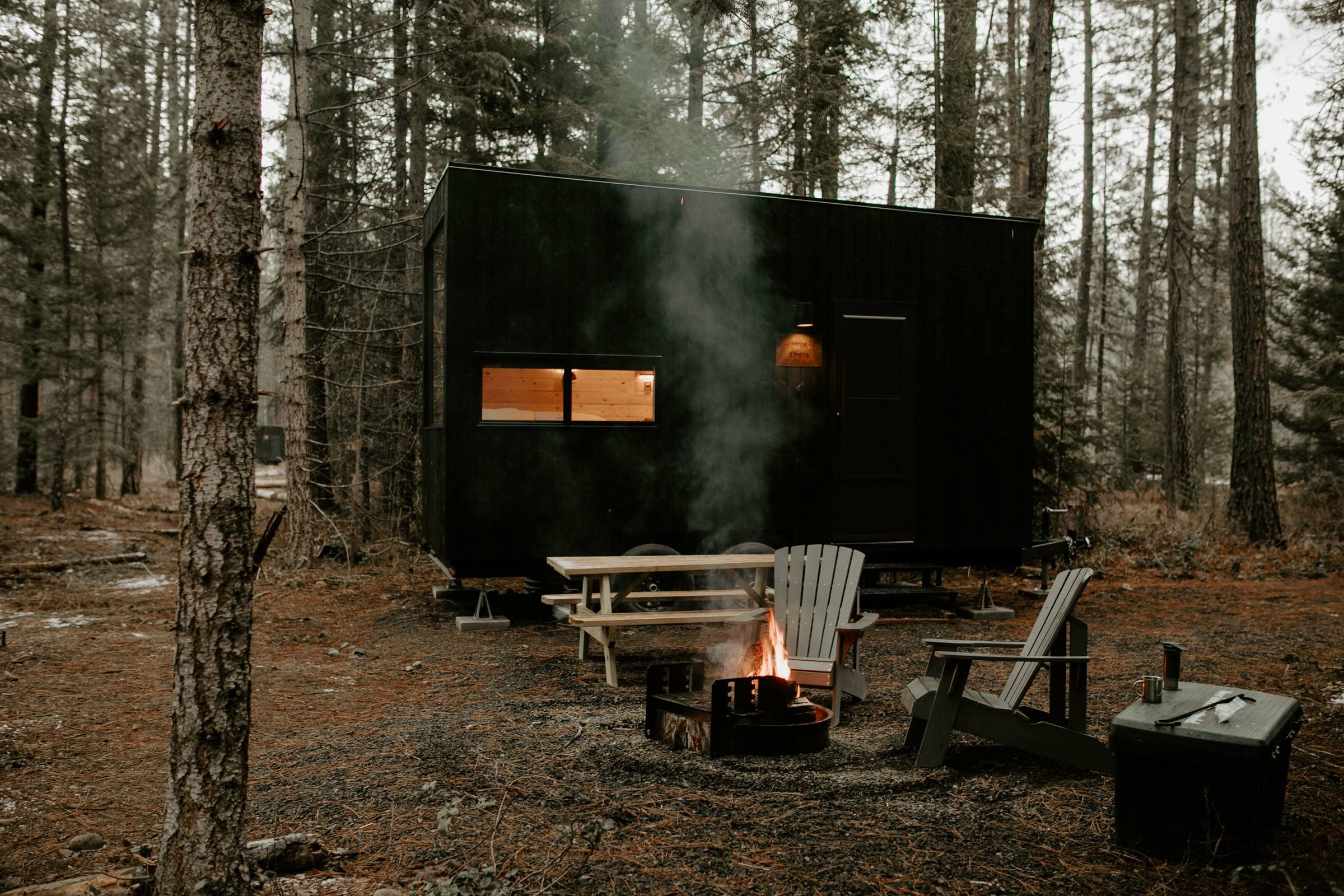 A small cabin on stilts with a black exterior, a picnic table, and two chairs around a fire pit in the middle of a wooded area