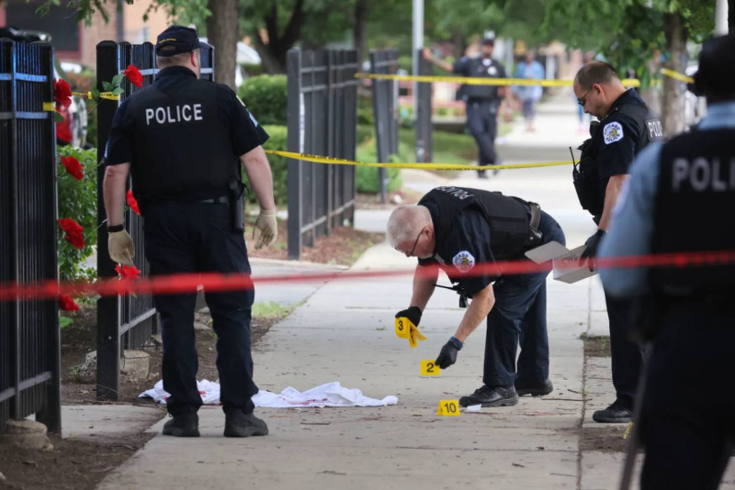 Police officers document a crime scene on a sidewalk with numbered evidence markers, yellow caution tape, and red flowers visible on a nearby fence.