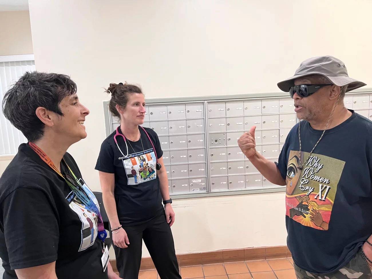 Two nurses talking to a person in a hat and sunglassses standing in front of a wall of metal mailboxes