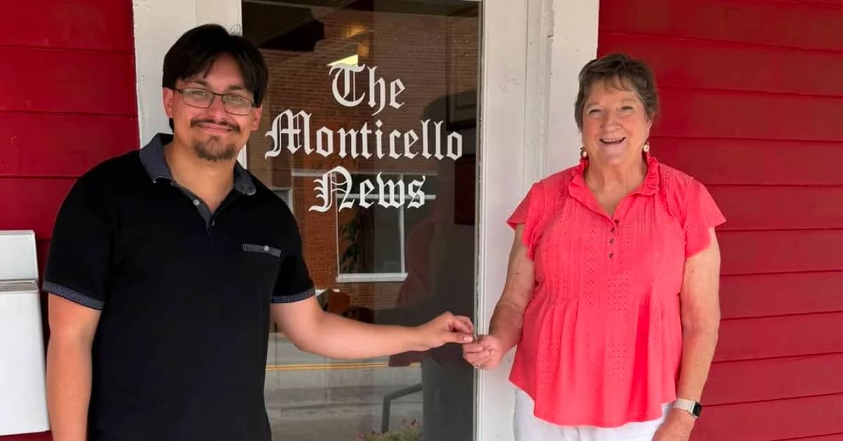 A woman in a red top handing a key to a man in a black polo, they're smiling and standing in front of a door that says "The Monticello News"