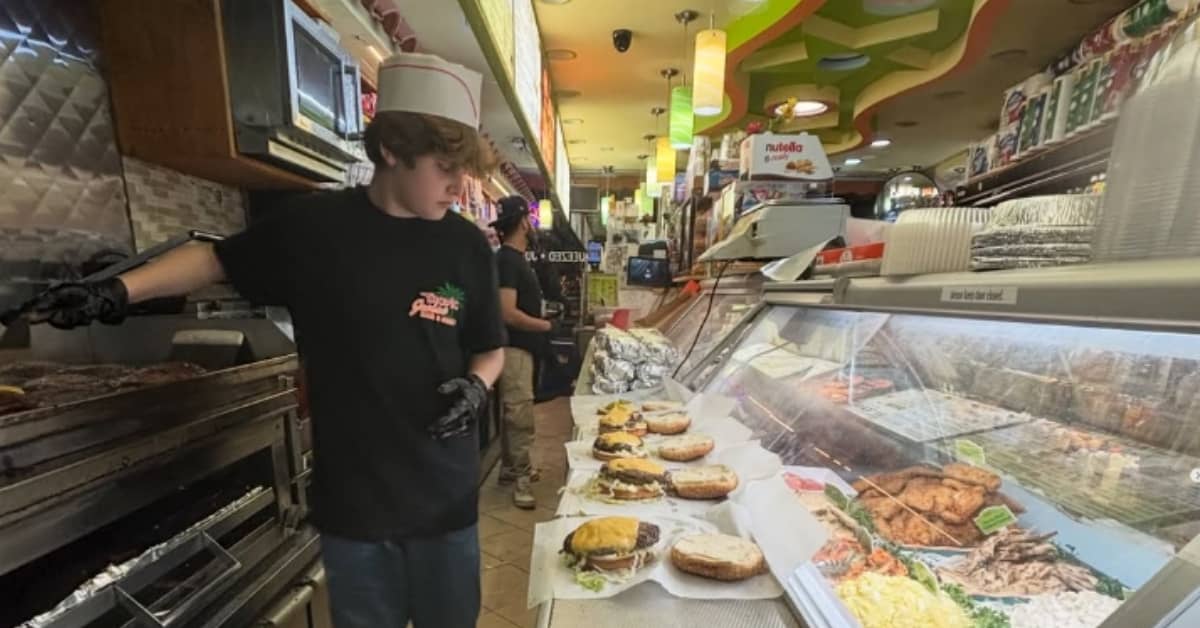 A young person behind a deli counter, wearing black tee, chef hat, and black gloves