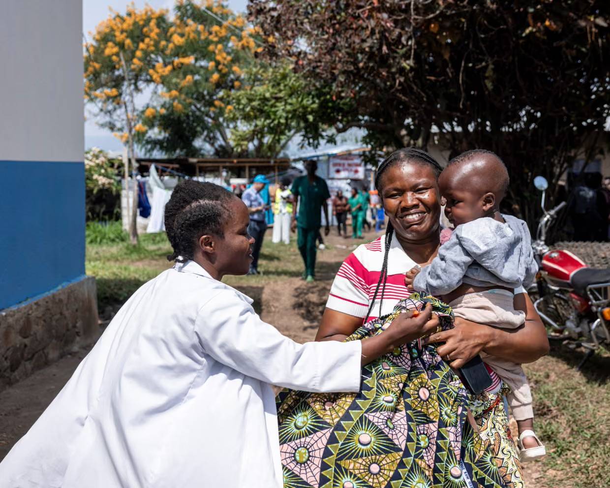 A person in a white coat reaching out to a woman holding their small child