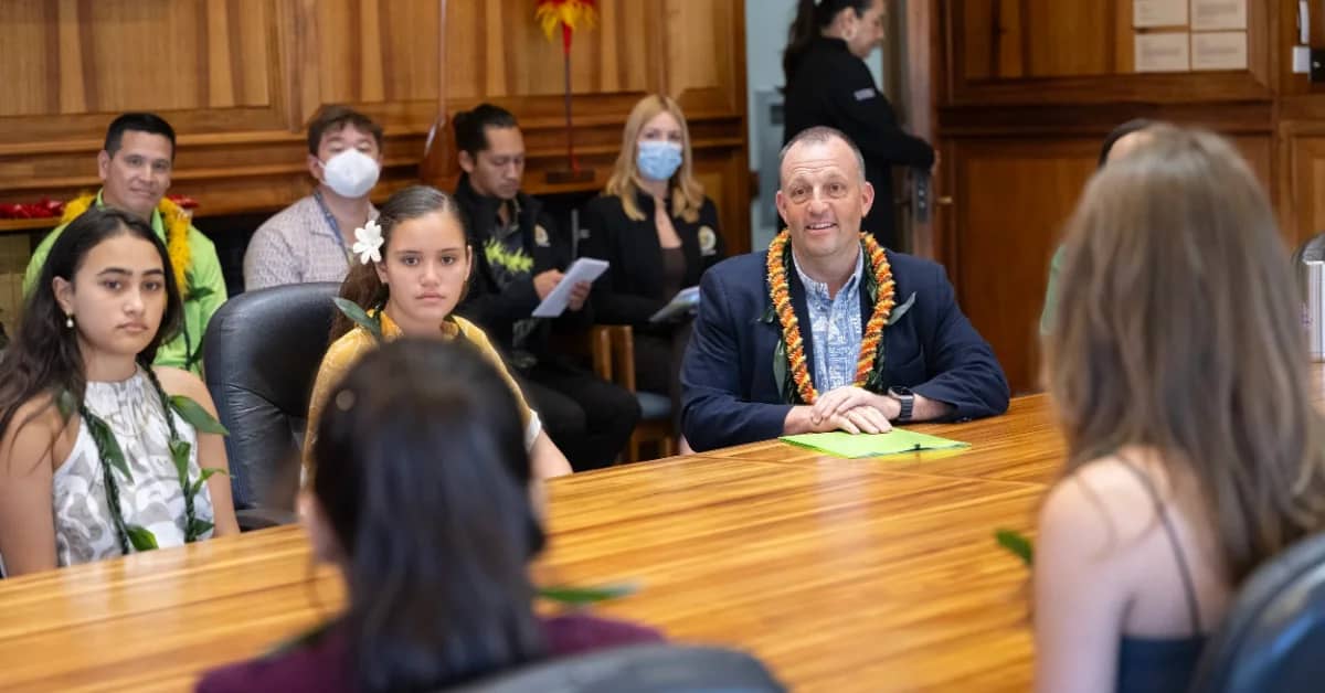 A man in a navy blazer and lei sits at a wooden table across from two women, with several other people wearing leis visible in the background of a wood-paneled room.