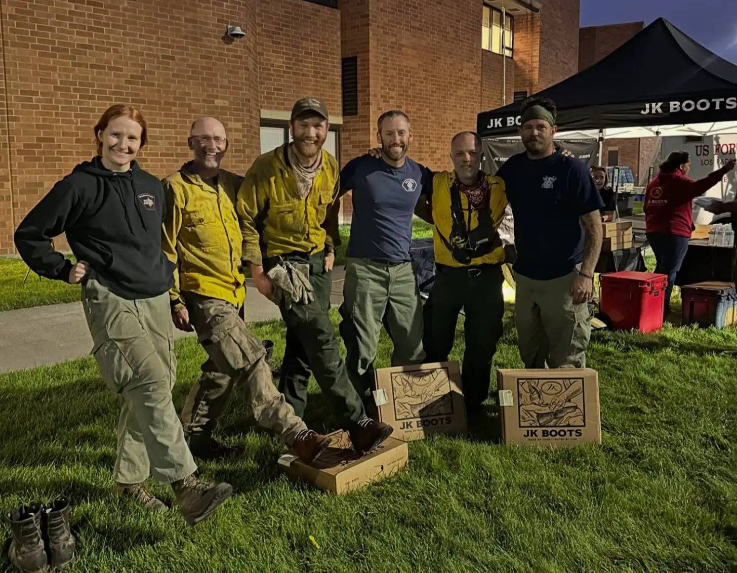 Six people stand on grass in front of a brick building, posing next to two wooden boxes labeled "JK BOOTS." A black tent and red containers are visible in the background.
